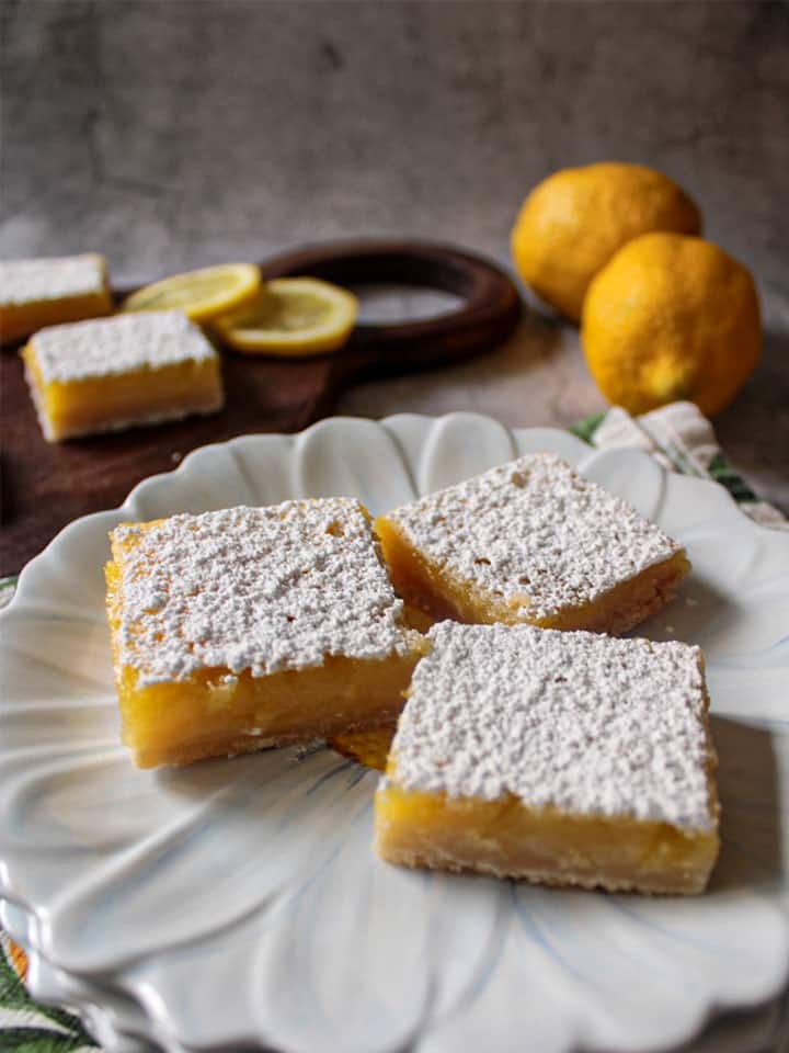 Lemon bars on a plate with more lemon bars on a platter in the background.
