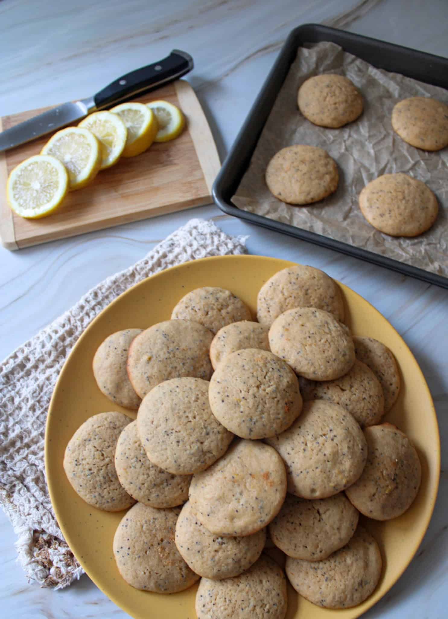 A plate of Lemon Poppy Seed Muffin Top Cookies with more on a cookie sheet in the background along with slices of lemon. 
